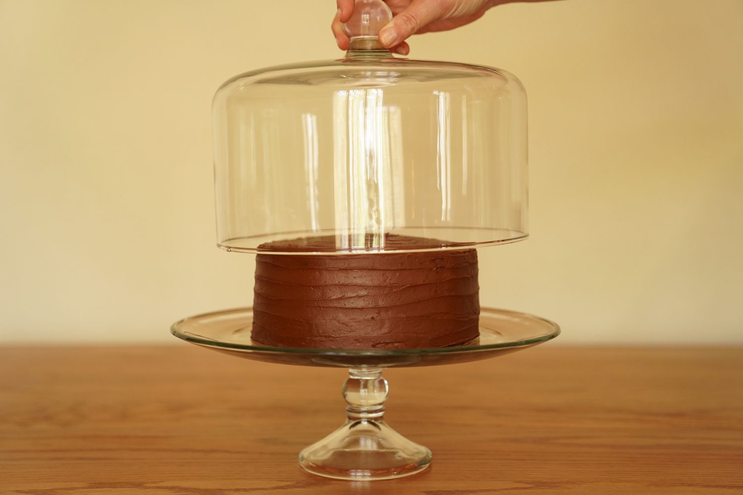 A glass cake stand holds a chocolate cake; a hand lifts a glass dome off the stand.