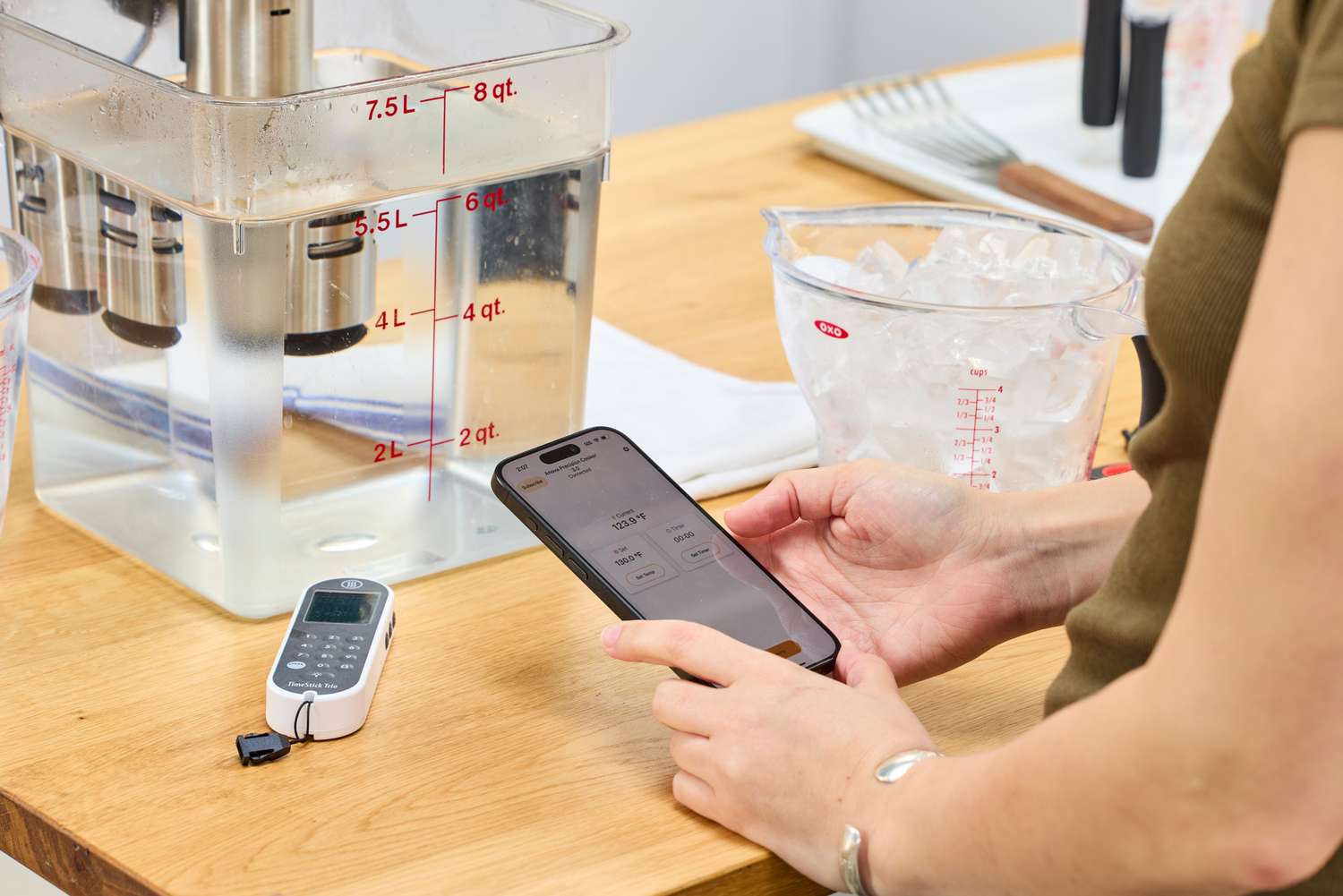 Person using a phone to control a Anova Culinary Precision Cooker 3.0 with a container of water and measuring tools on a nearby table
