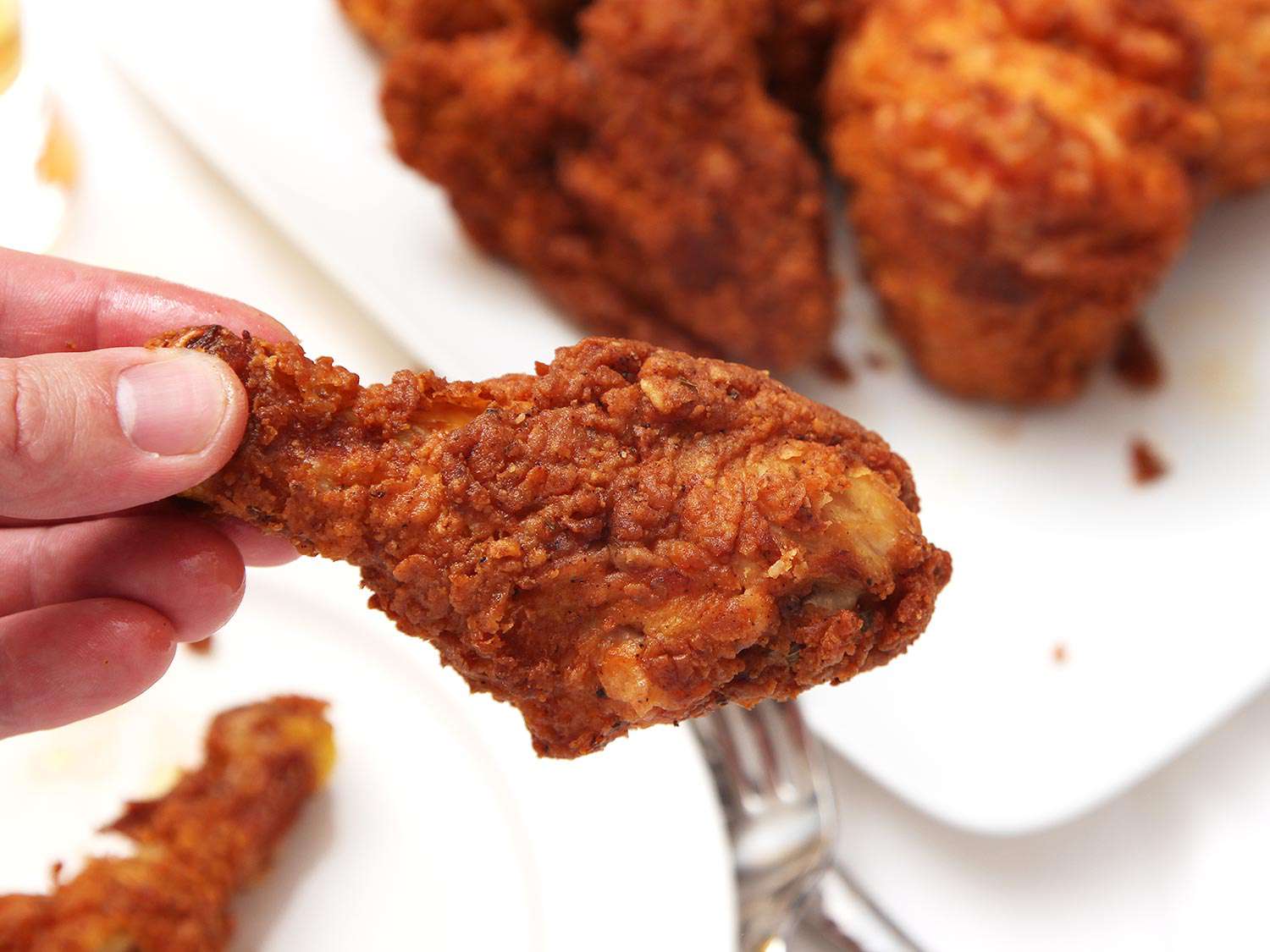 Hand holding a crispy fried chicken drumstick pierced with a fork.