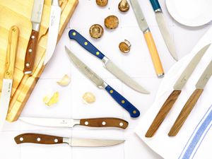 a variety of steak knives on a white tile surface with garlic cloves and mushrooms