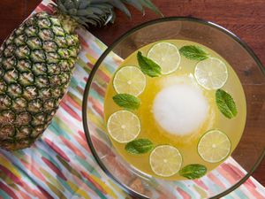 Overhead view of Isla Bonita Punch, garnished with mint leaves and lime wheels. A large sphere of ice floats in the center of the punch bowl. A whole pineapple rests on its side nearby.