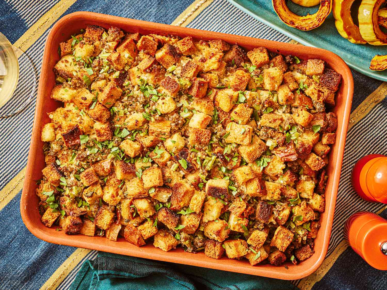 sausage and sage stuffing in orange baking dish, on a thanksgiving table, with roasted squash to the side. 