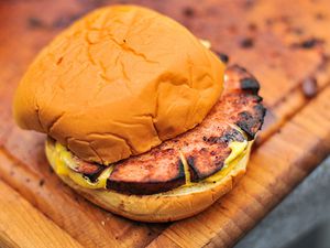 A slab of crisp-edged smoked bologna sandwiched in a sauced bun, sitting on a cutting board.