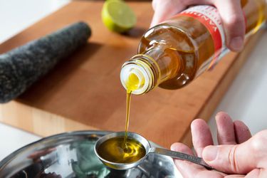 Mustard oil being poured into a measuring spoon. A cutting board, knife, pestle, and half a lime are out of focus in the background.