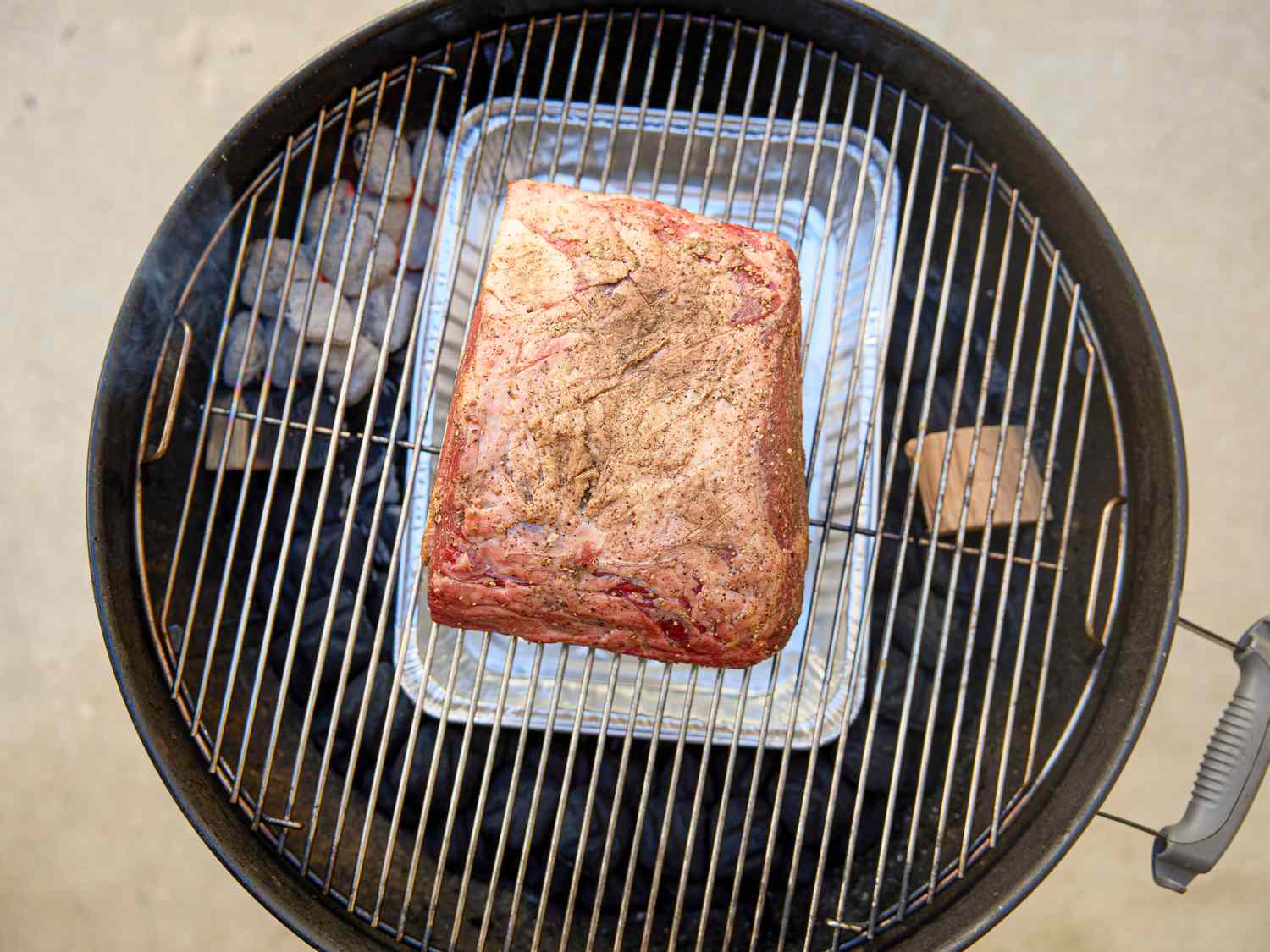A prime rib being smoked on a circular grill with coals and a wood block on one side