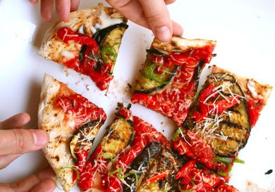 A grilled pizza, sliced into pieces and being taken by several hands on a white background.