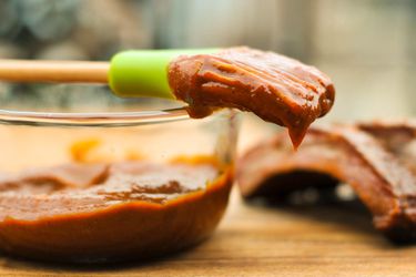 A glass bowl of Kansas City-style barbecue sauce with a basting brush dipped in sauce on top of the bowl. Some cut ribs are in the background. 