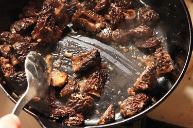 Sautéed morel mushrooms in a cast iron skillet being stirred with a large slotted spoon. 