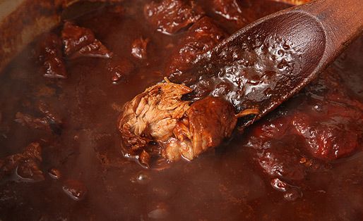 Stirring carne adovada in a pot with a wooden spoon; the spoon is lifting up a piece of cooked pork that's been torn in half. 
