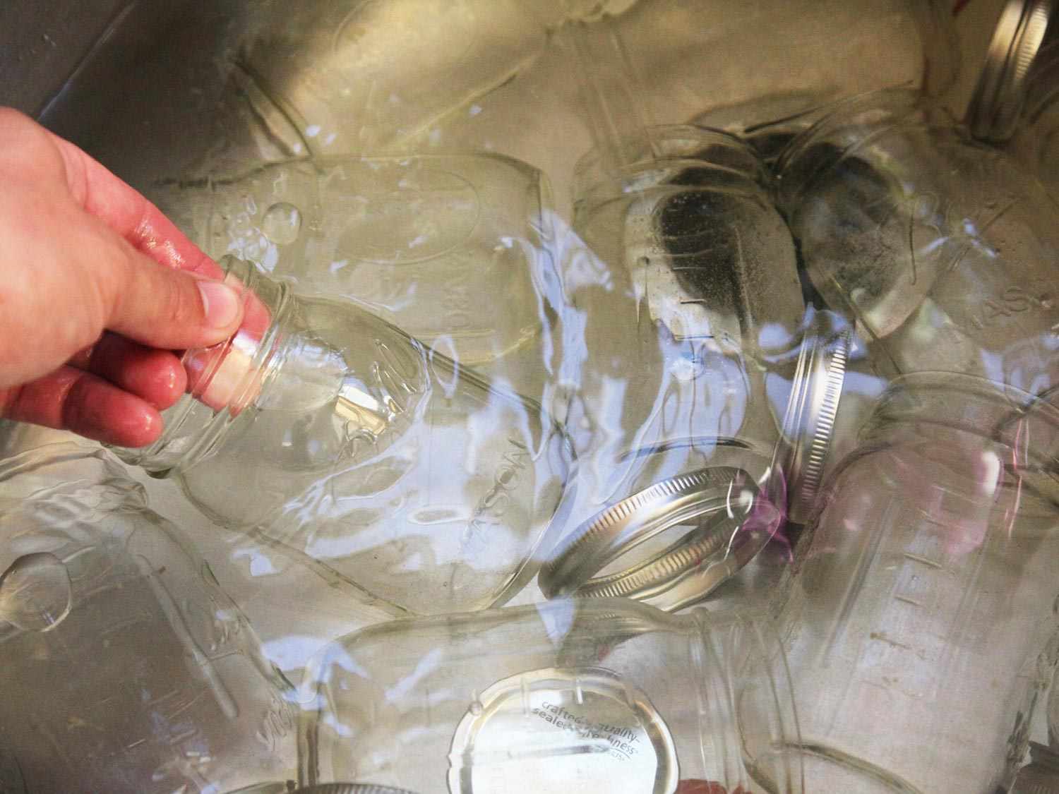 A hand lifting a glass canning jar out of a sink full of water holding many glass jars, metal lids, and metal rings.