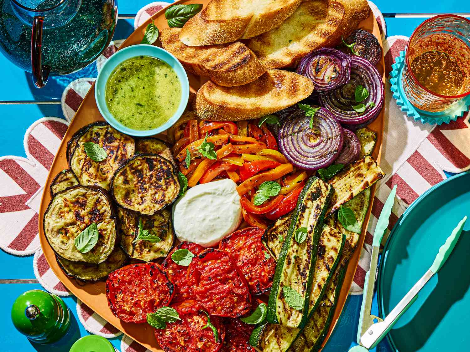 An assortment of grilled vegetables including zucchini, eggplant, onions, and tomatoes, served with bread and dips on a platter