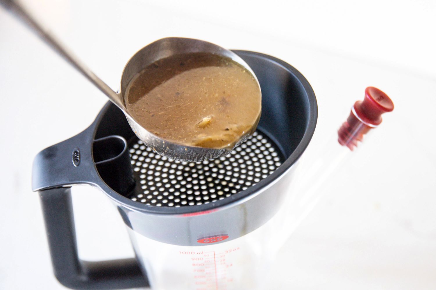 Metal ladle pouring broth through a mesh strainer placed over a measuring container