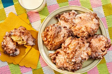 Platter of apple fritters on a round dish, with one fritter on yellow napkins, ripped open. Glass of milk, and colorful checkered tablecloth