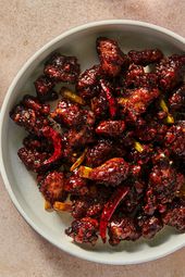 A wide ceramic bowl holding General Tso's Chicken. In the upper left corner of the image is a ceramic bowl of white rice, and in the top right corner is a glass holding a beverage. There is another bowl in the bottom left corner.