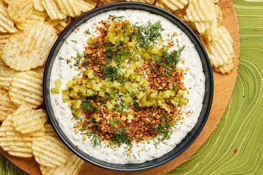 Bowl of pickle dip garnished with panko, dill and pickles, surrounded by a wooden plate full of potato chips, on a green tabeltop