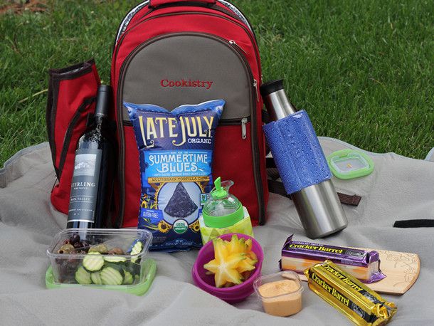 Chips, pickles, cut fruit, cheese, and dips around a backpack picnic basket on a gray picnic blanket spread over grass. 