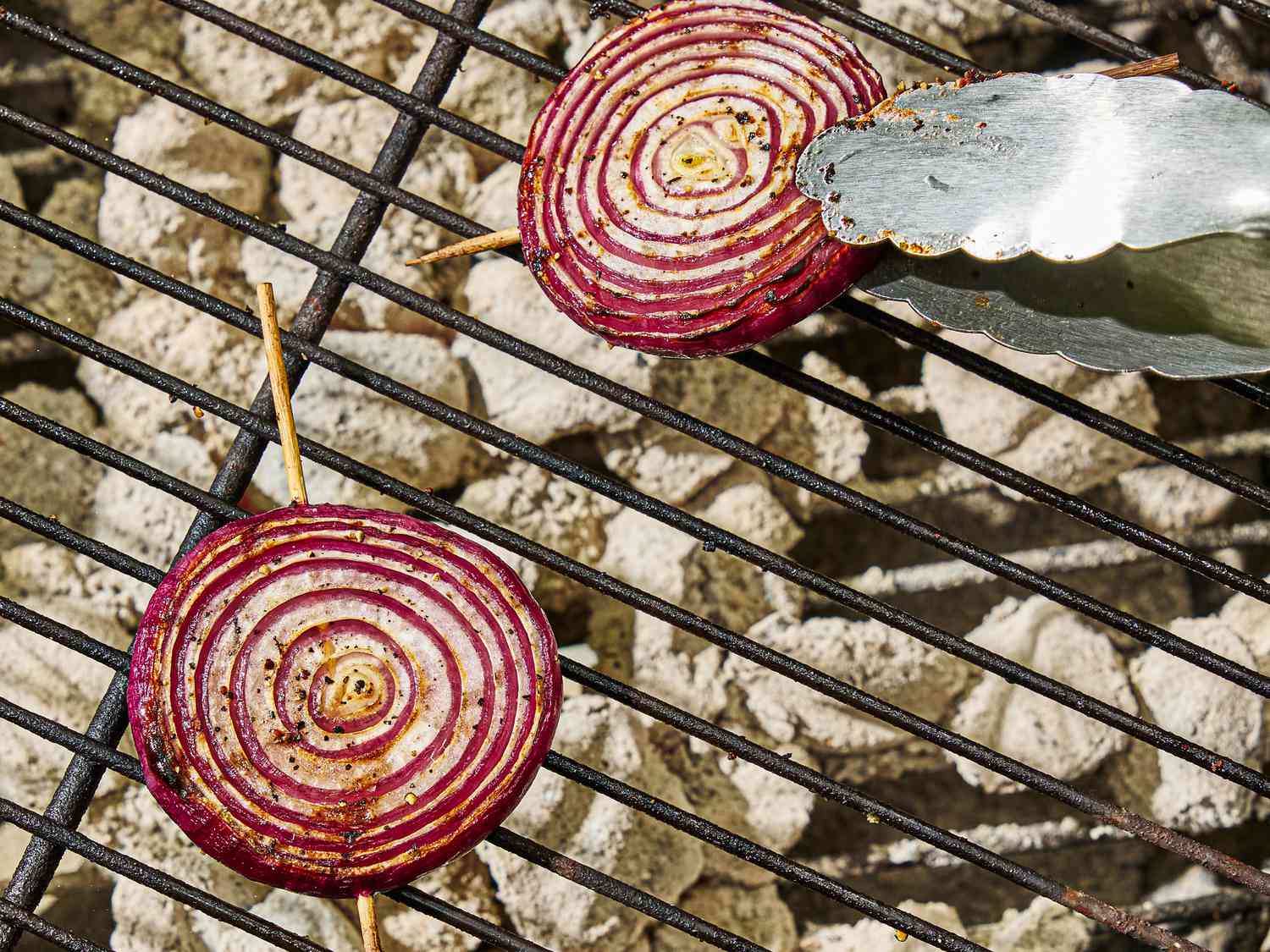 Slices of onion on a skewer being grilled over a charcoal barbecue, with tongs turning one of them