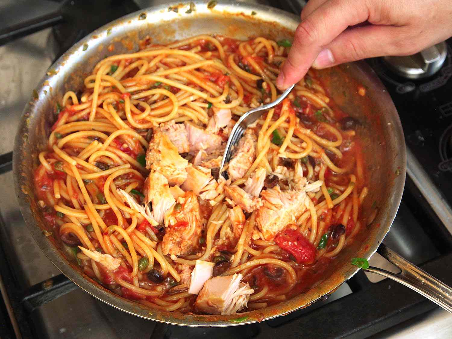 Adding canned tuna to a skillet of pasta puttanesca.