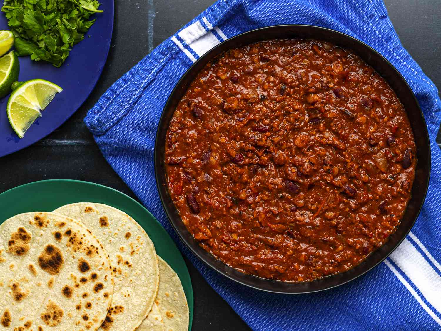 A large bowl of chili on a blue dish towel. To the bottom left of the image is a plate holding tortillas, and in the top left corner is a plate with chopped cilantro and sliced limes.