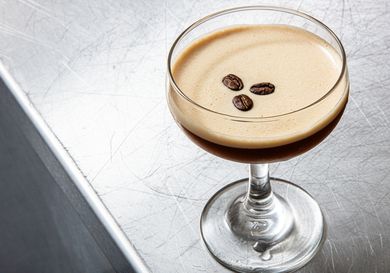 A glass of espresso martini garnished with three coffee beans on a metallic surface