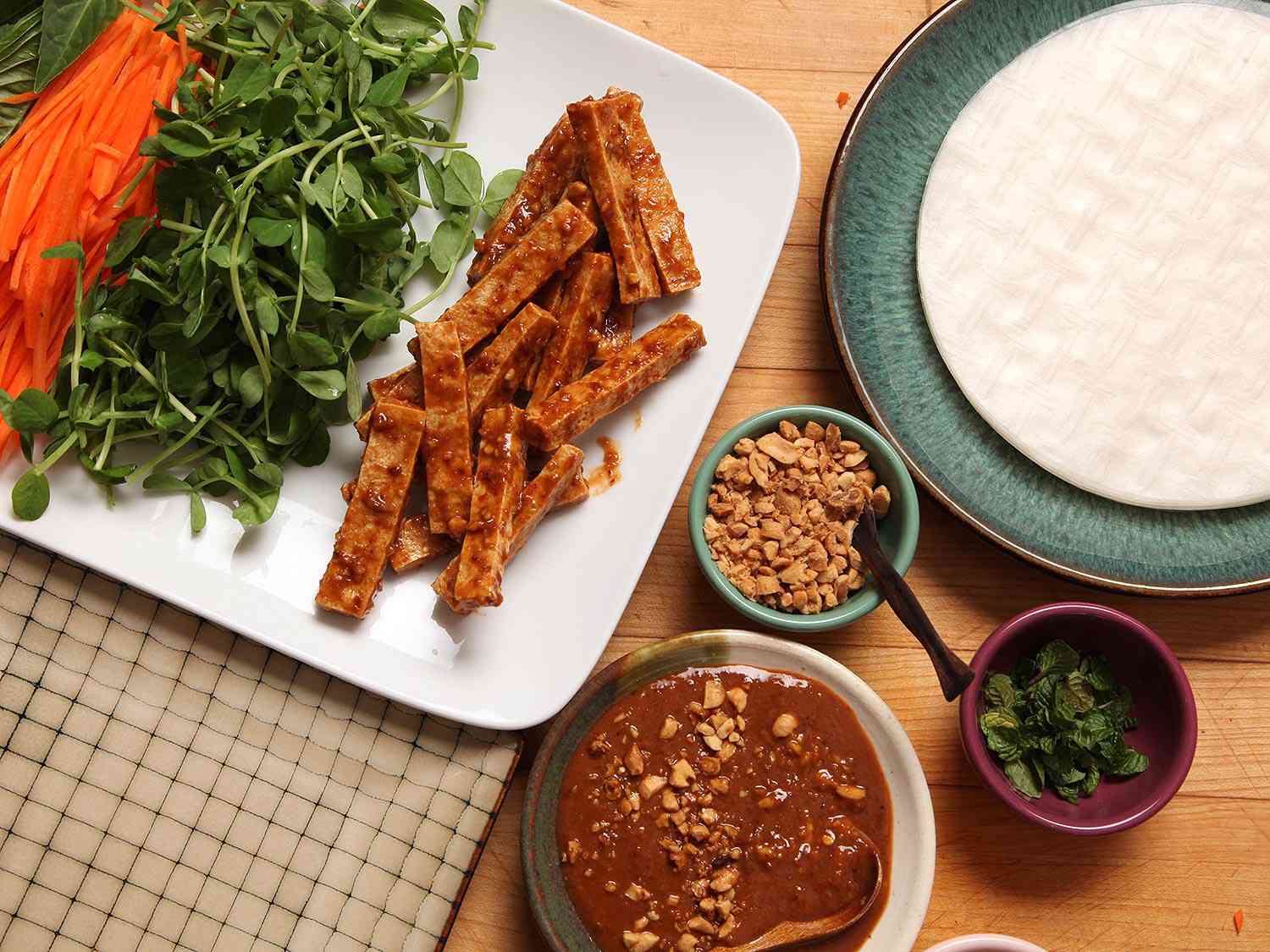 Overhead view of all the fillings for the vegan spring rolls are arrayed next to a stack of rice paper rounds, the dipping sauce, and small ramekins containing crushed peanut and chopped cilantro.