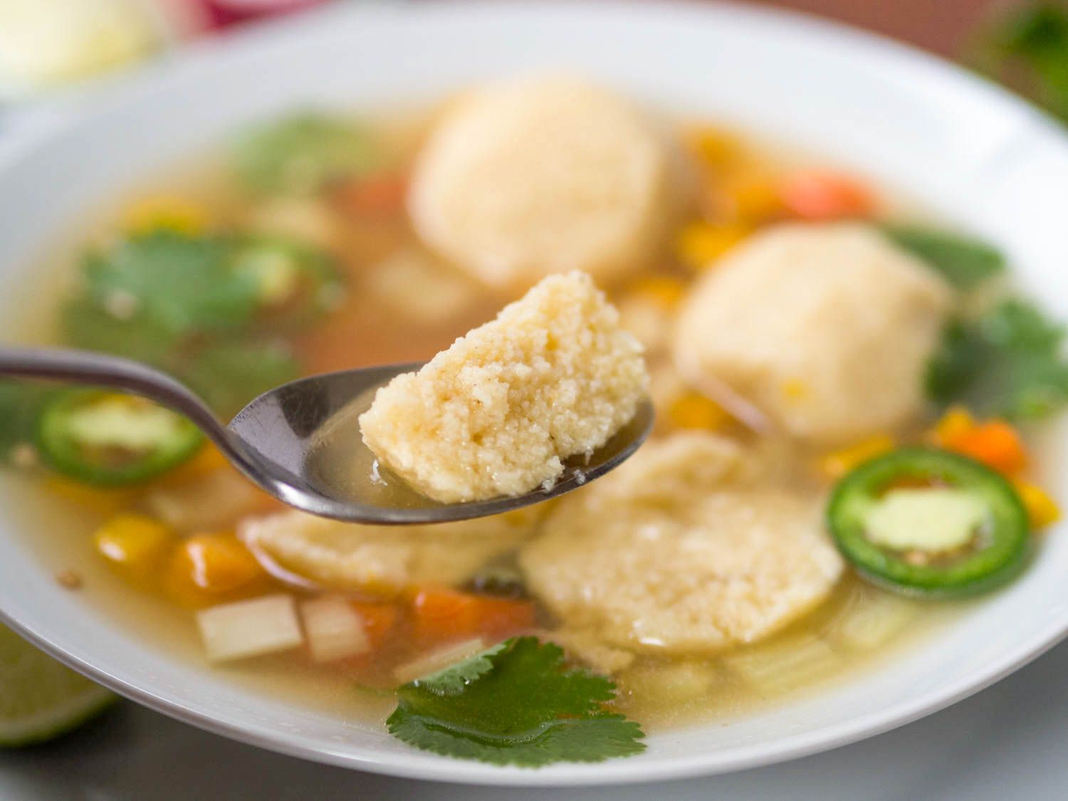 Closeup of a spoon with part of a masa-ball on it held above a bowl of masa-ball soup.
