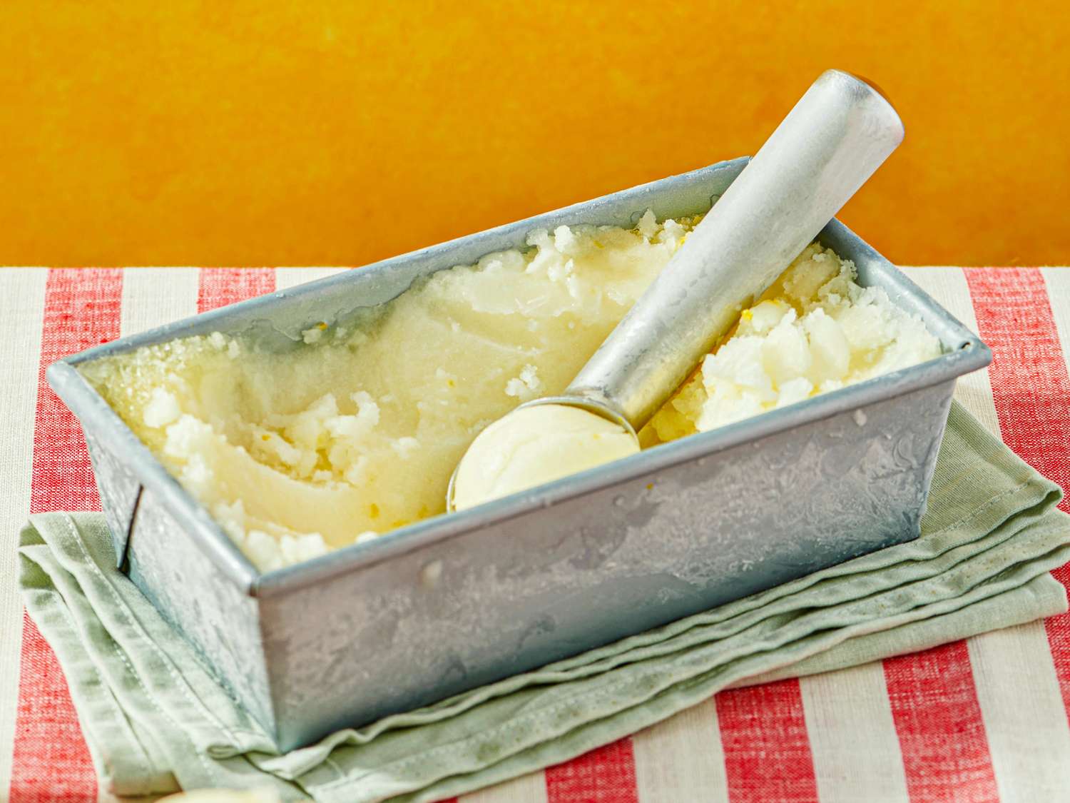 Metal container holding lemonflavored shaved ice with a scooper on top placed on a striped towel