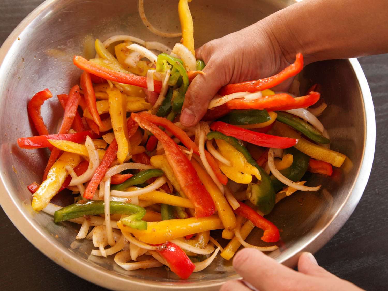 Hands tossing strips of onion and green, yellow, and red bell pepper in a metal bowl for fajitas.