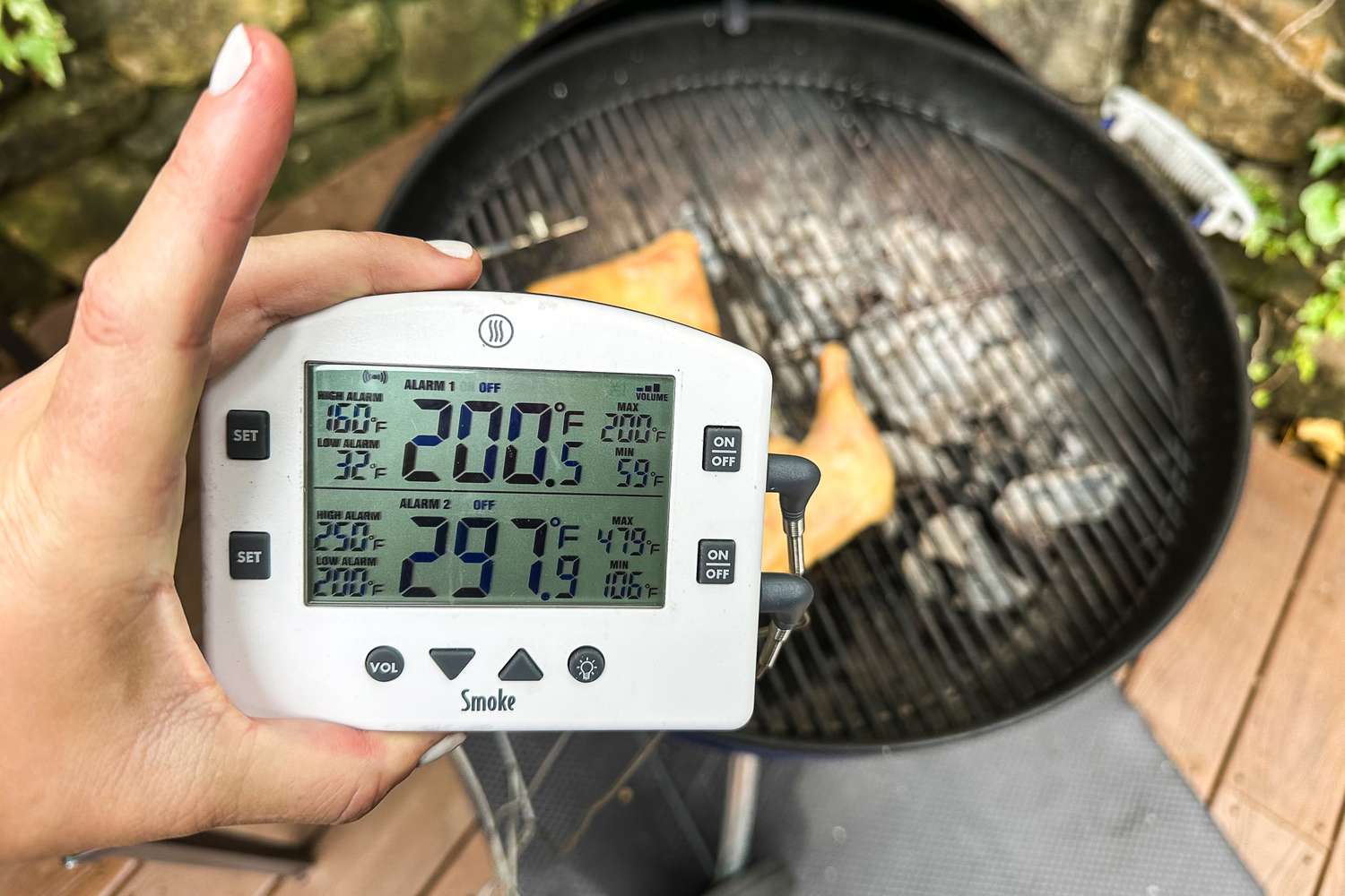 A hand holding a thermometer displaying temperatures with a barbecue grill and food visible in the background
