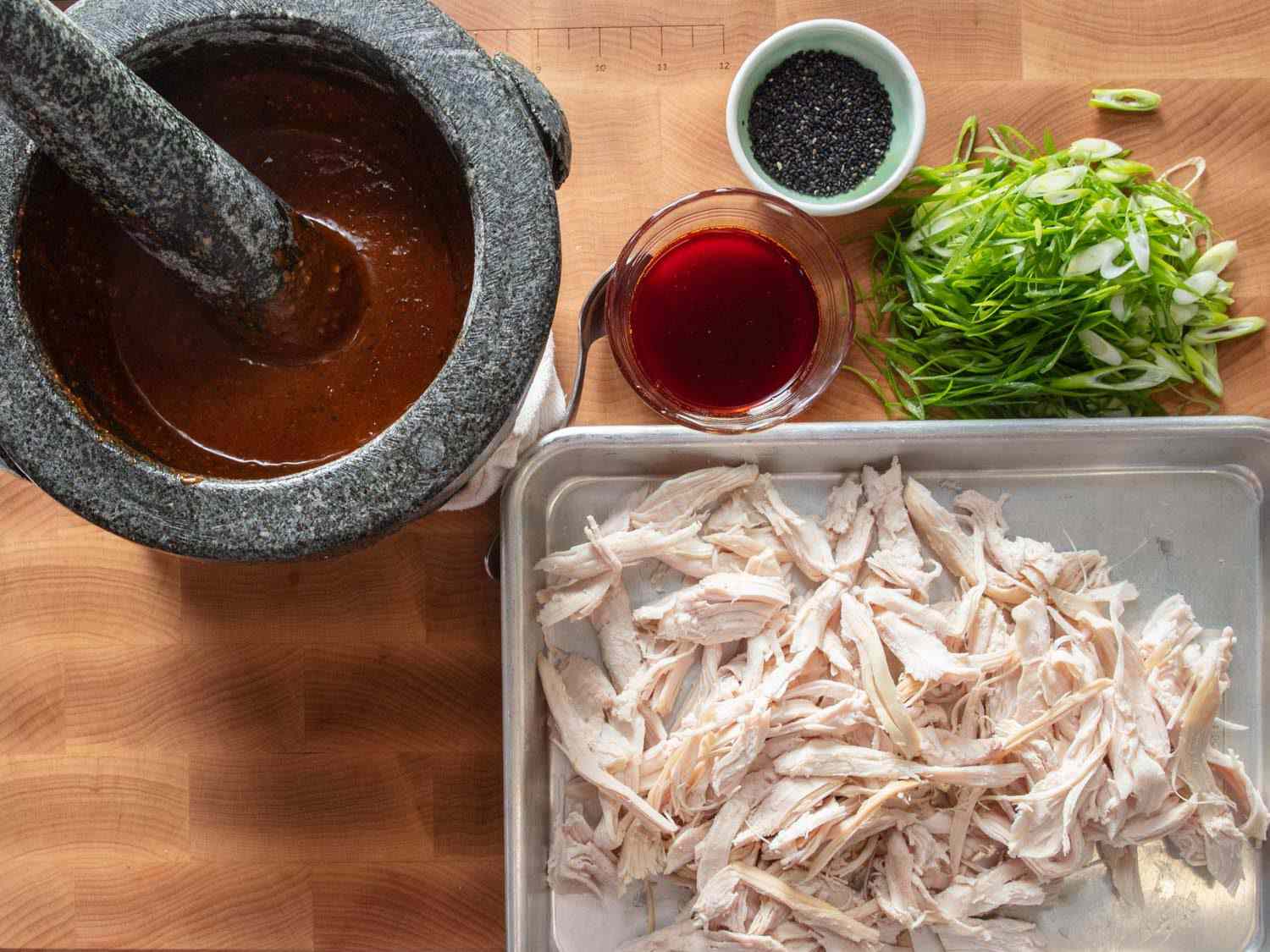 Overhead of components for Sichuan-style turkey salad: dressing in a mortar and pestle, pulled roast turkey breast meat, chili oil, sliced scallions, and black sesame seeds.