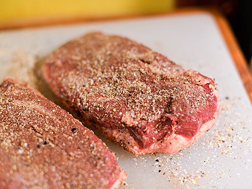 Two duck breasts covered in salt and spices on a plastic cutting board. 