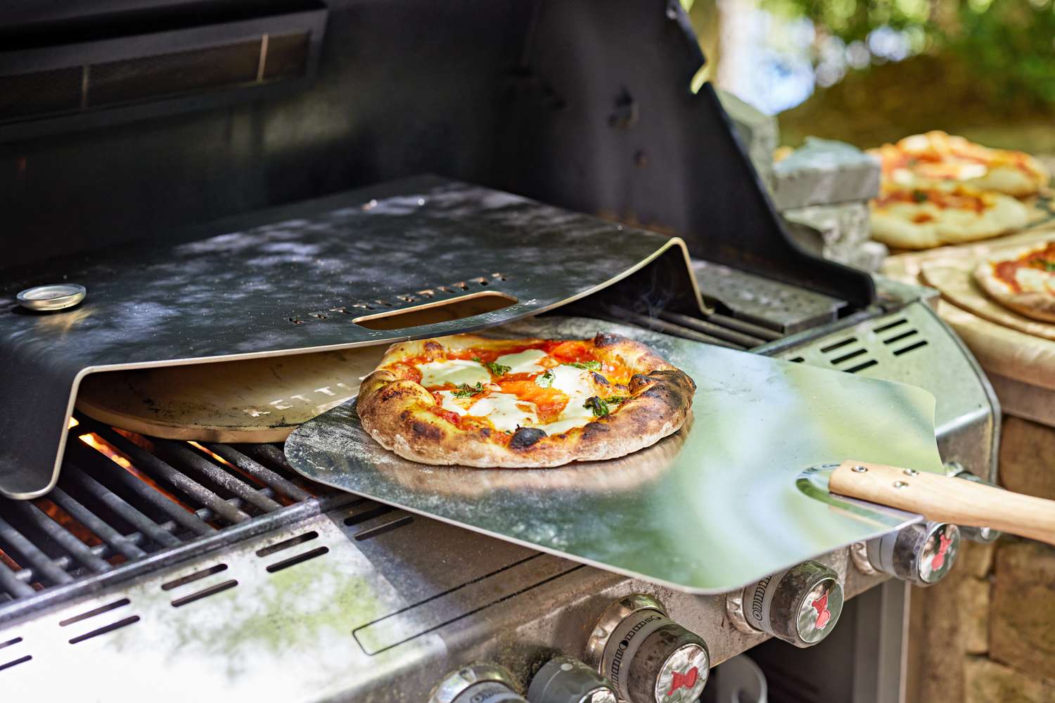 Pizza being removed from between pizza stone and stainless steel lid of KettlePizza Pro Pizza Oven Kit set on gas grill 