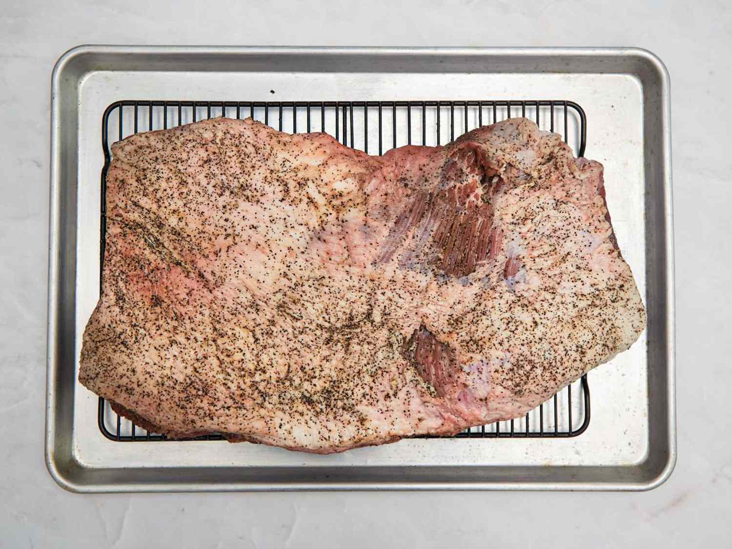 A seasoned brisket on a wire rack placed on a baking sheet, ready for smoking preparation