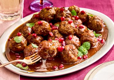 Plate of cocktail meatballs garnished with pomegranate seeds and mint leaves set on a tablecloth with glasses in the background