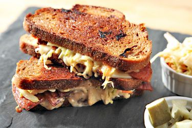 Two Rachel sandwiches stacked on top of each other, on a slate cheese board with containers of pickles and slaw