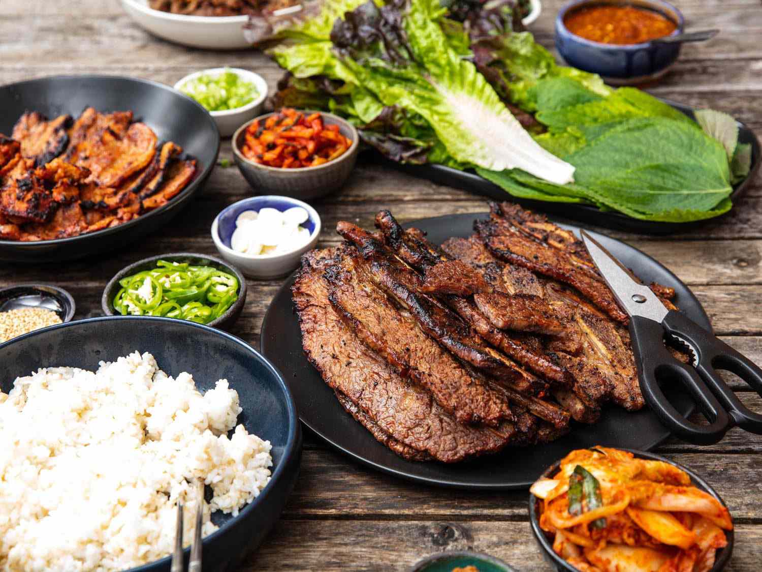 Side view of a table set with a Korean barbecue spread, with a plate of beef galbi in the foreground.