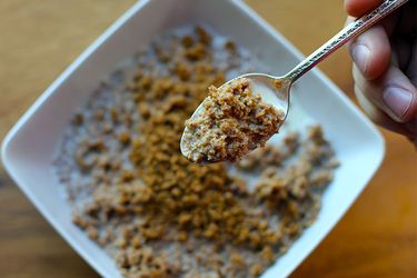 Overhead view of a spoonful of DIY grape-nuts cereal. A bowl of the cereal is visible in the blurred backgroung.