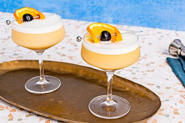 Two whiskey sour cocktails, garnished with an orange wheel and a cherry on a cocktail pick, served on a tray