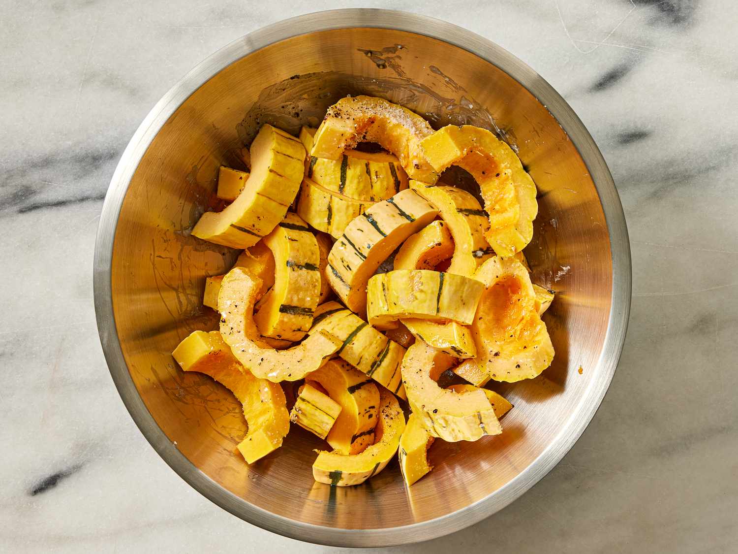 Squash in a metal bowl, tossed with seasonings, on a marble surface 