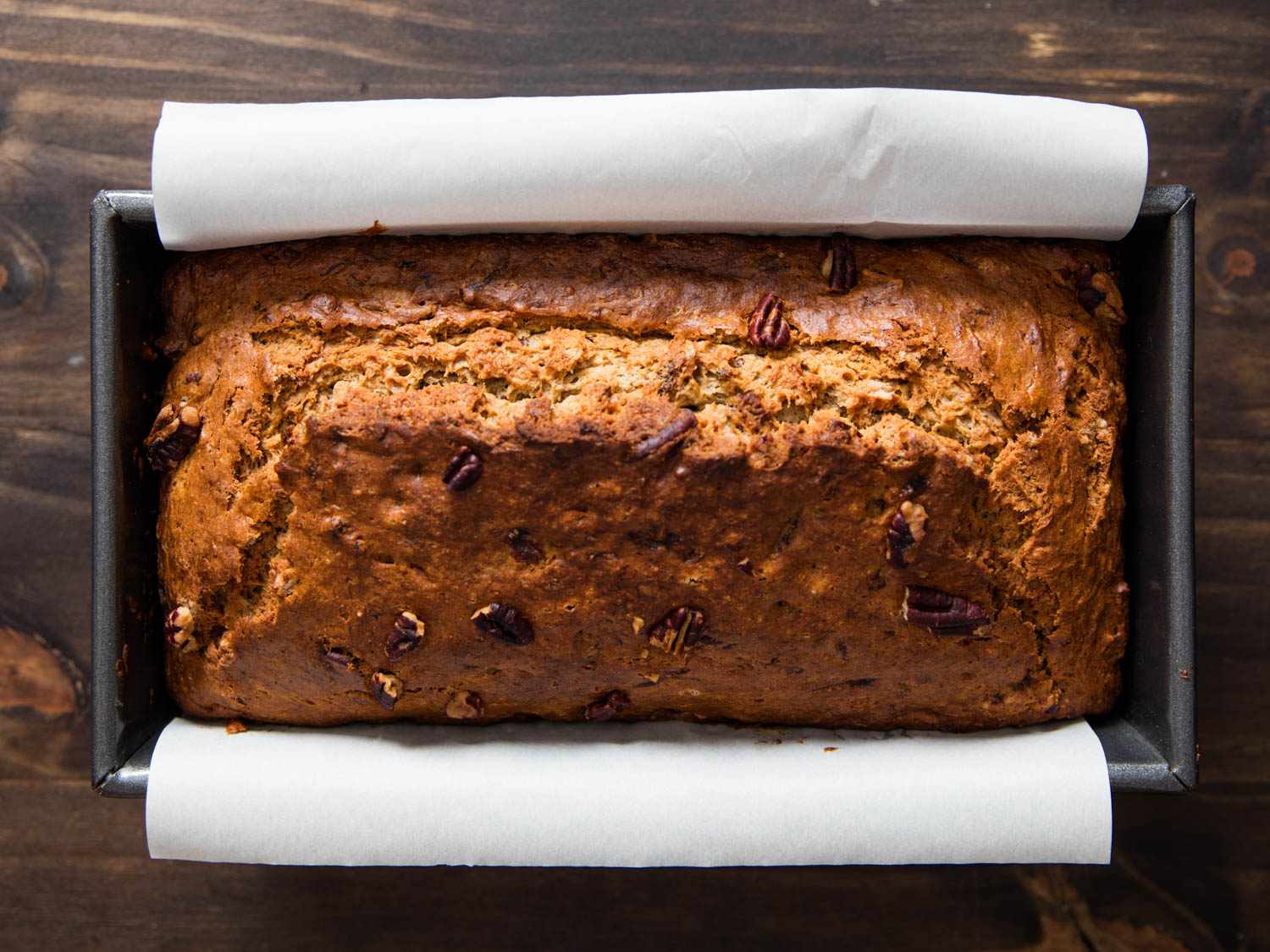 Baked banana bread in a parchment-lined loaf pan on a wooden cutting board.