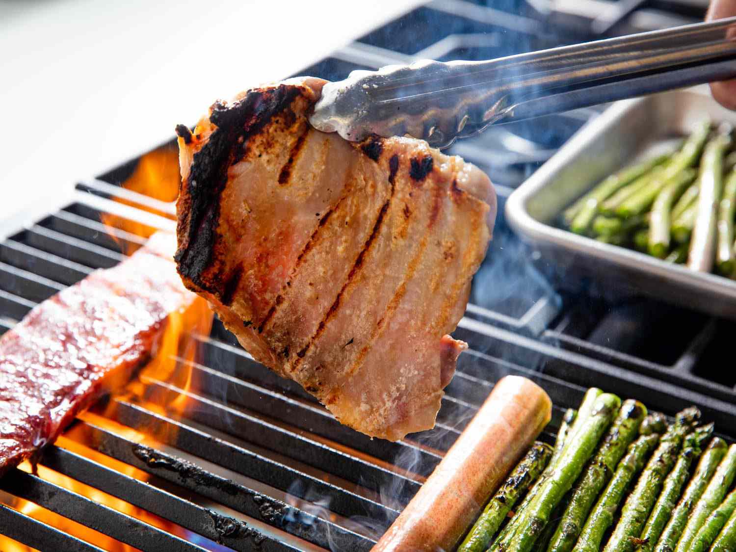 A chicken thigh on a standard gas grill grate held aloft by tongs shows uneven browning, with one stretch of severe char along one edge and not enough browning on the opposite one; flames flare up in the background, meanwhile