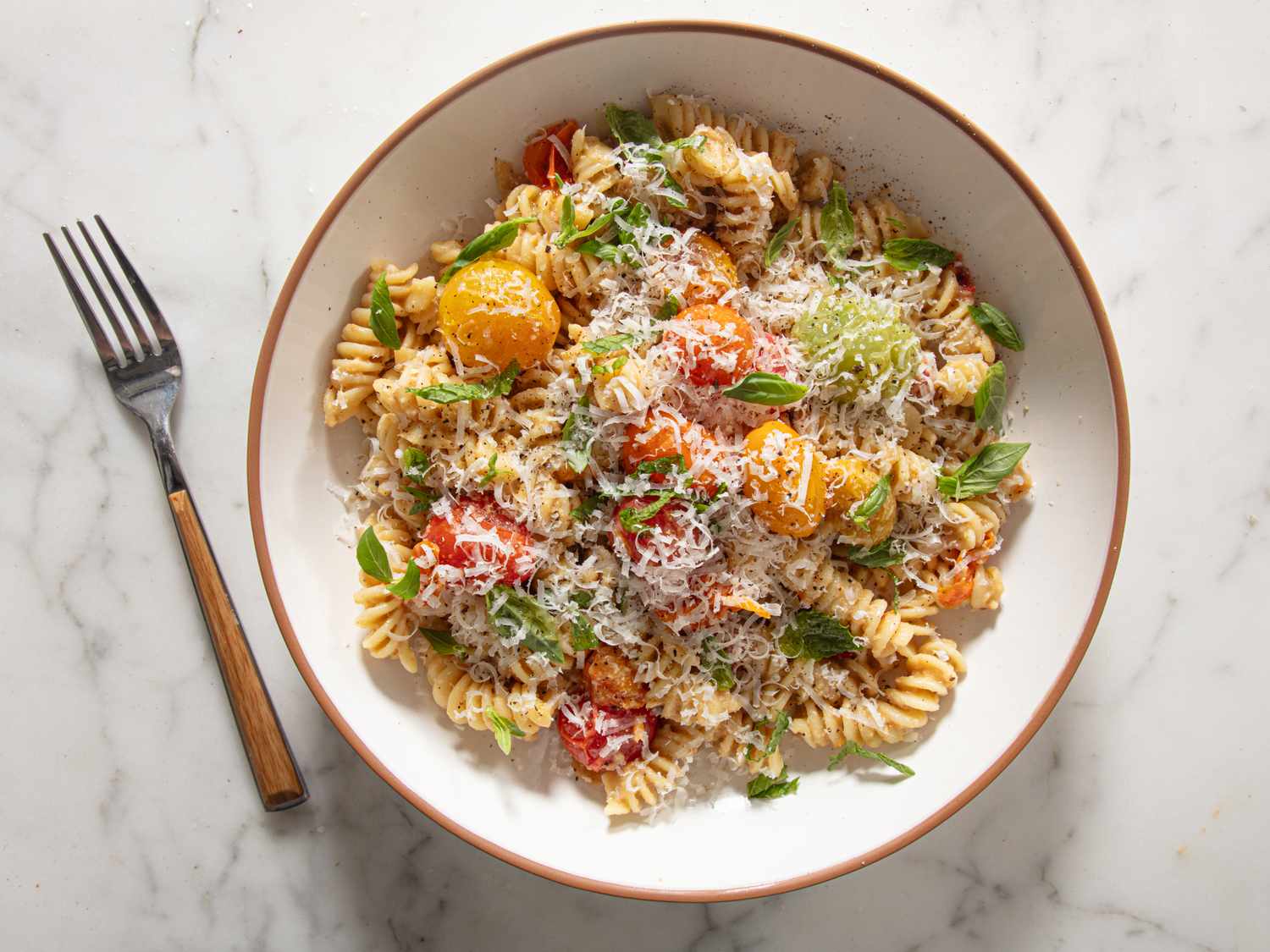 A bowl of pasta garnished with cherry tomatoes herbs and grated cheese presented next to a fork on a marble surface