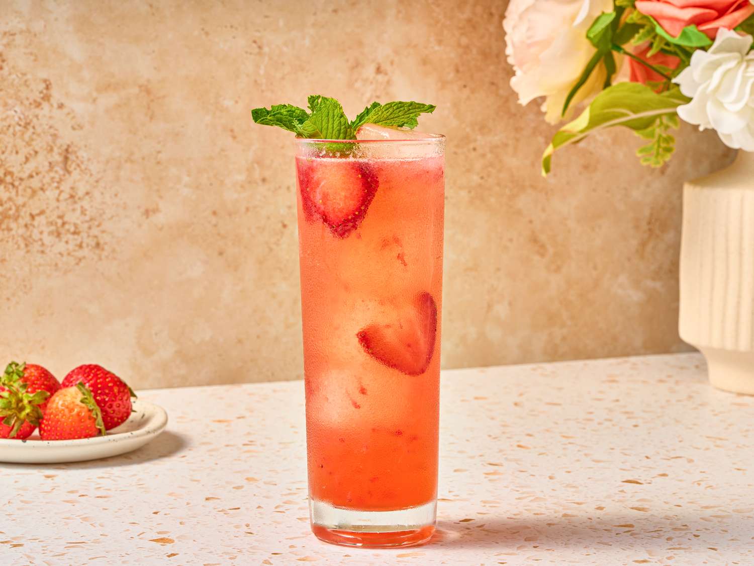 A strawberry mint sparkler served in a glass with mint garnish set on a table near flowers and fresh strawberries