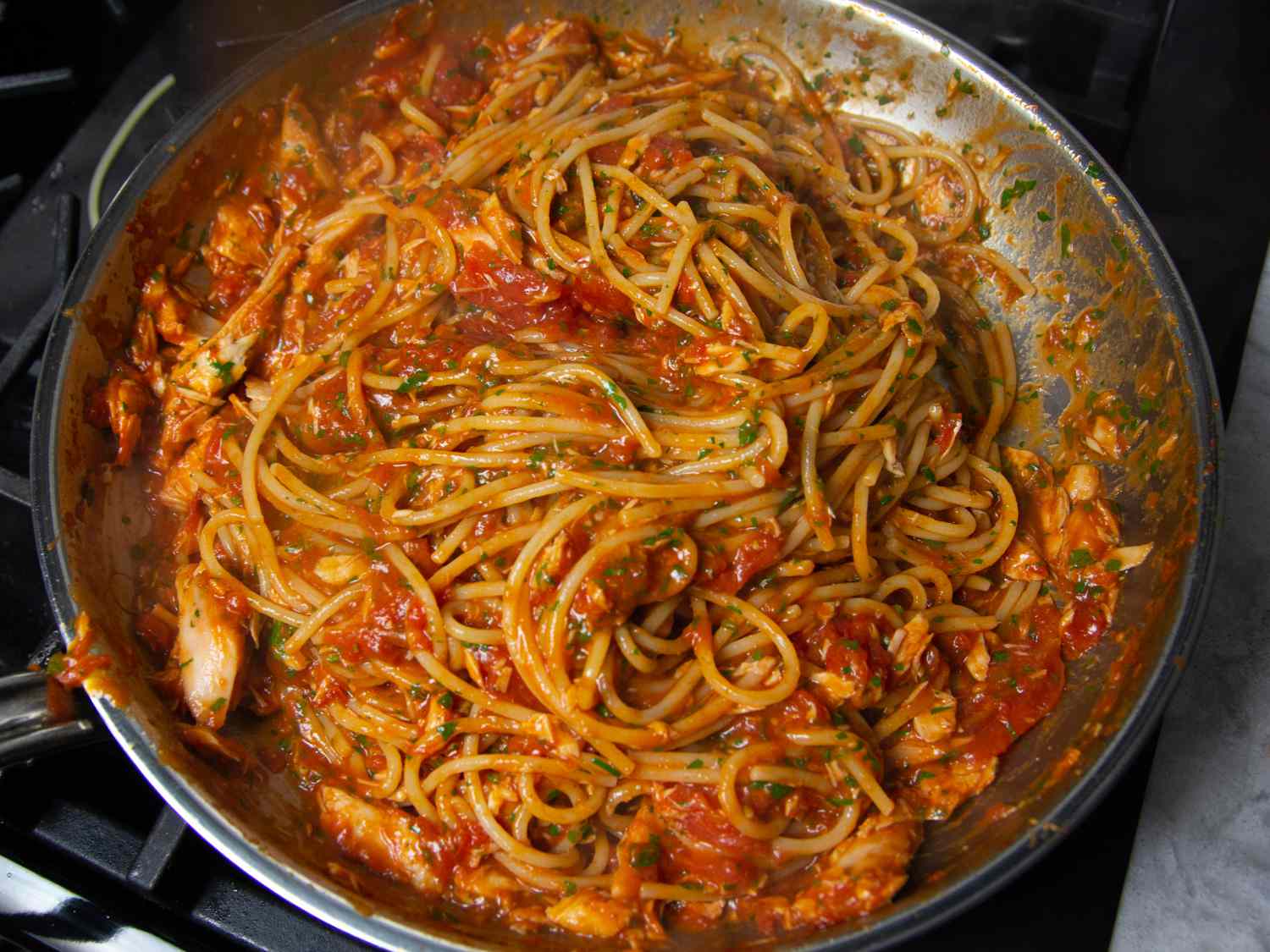 A skillet with spaghetti and tomato-based sauce, featuring visible parsley and pieces of tuna