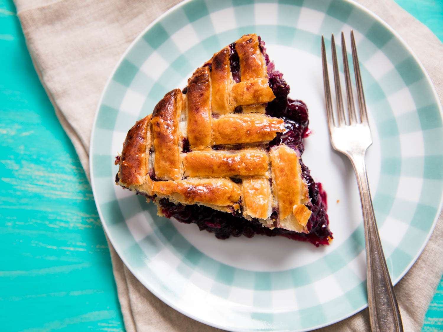 A wedge of herringbone lattice–topped blueberry pie on a plaid-rimmed plate with a fork