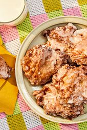 Platter of apple fritters on a round dish, with one fritter on yellow napkins, ripped open. Glass of milk, and colorful checkered tablecloth