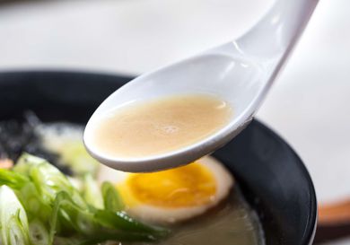 Closeup of a spoonful of chicken paitan broth. A bowl of ramen is visible in the blurred background.