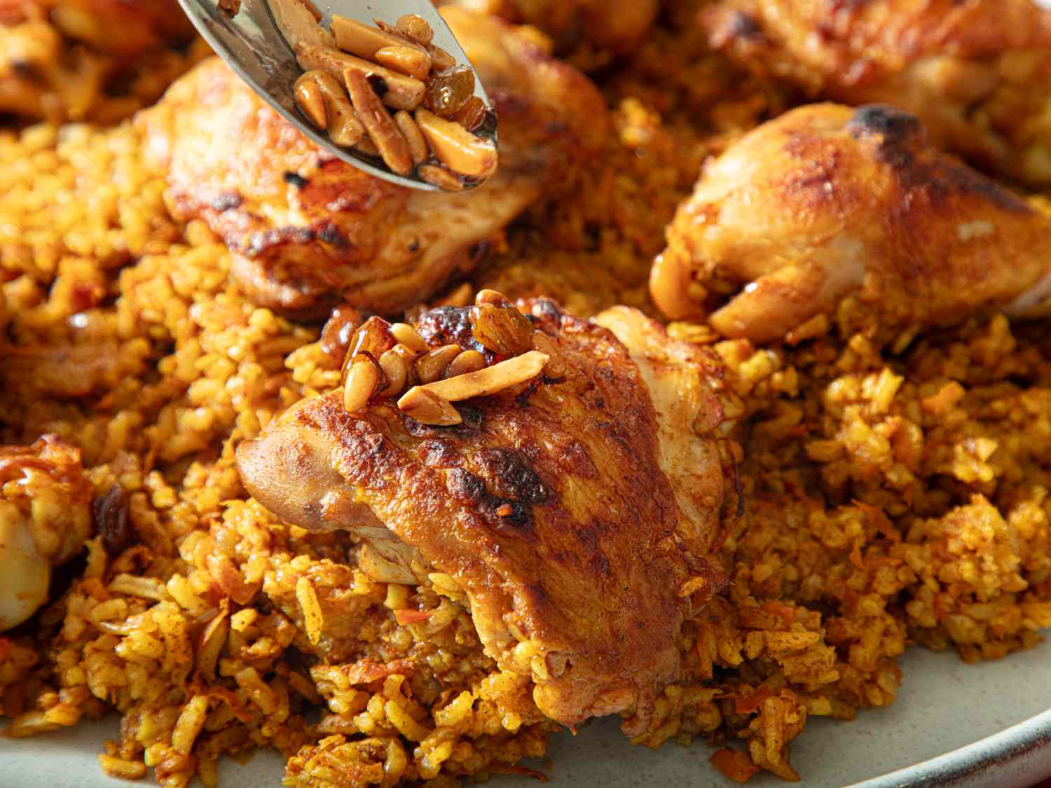 A dish of kabsa with roasted chicken pieces and a spoon scooping toasted almonds on top