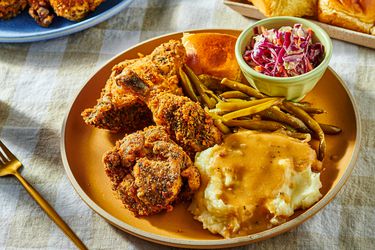 Dish of fried chicken with coleslaw, greenbeans, and mashed potatoes, with a fork and serving dishes in the background