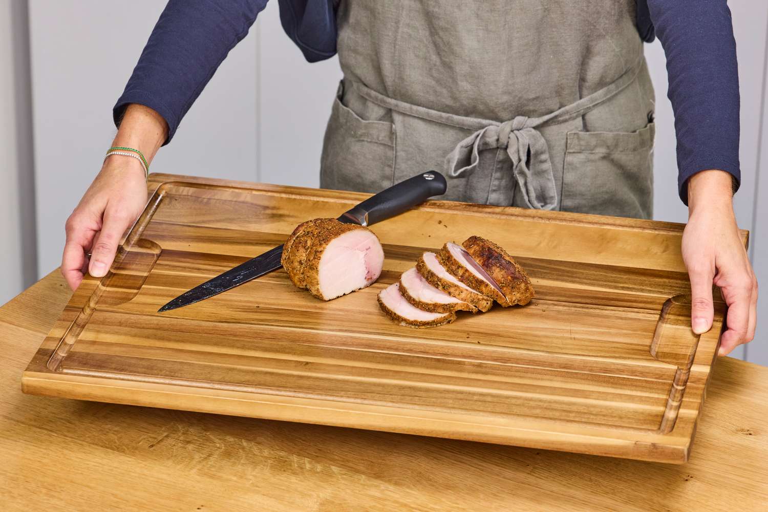 A person setting down an acacia wood cutting board with a sliced pork tenderloin on top of it.